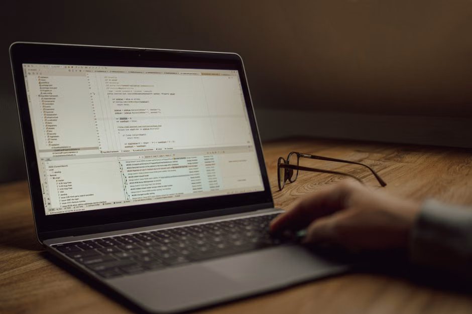 Pessoa trabalhando em código de programação em um laptop dentro de casa. Óculos sobre a mesa.
